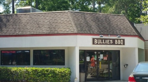 Exterior of Bullies BBQ restaurant with a sloped roof and large windows, surrounded by greenery.