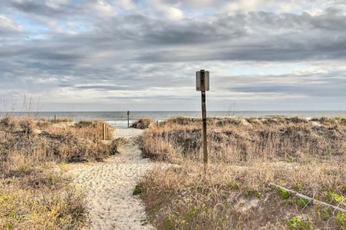A sandy path leads through tall grass to the ocean, under a cloudy sky with a signpost nearby.