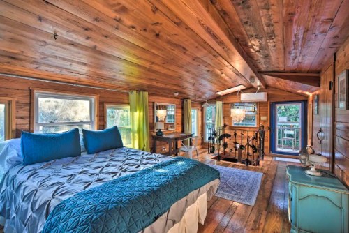 Cozy wooden bedroom with a blue quilted bedspread, large windows, and a small workspace in a rustic setting.