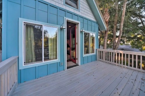 A blue house with a deck, featuring large windows and an open door, surrounded by trees.
