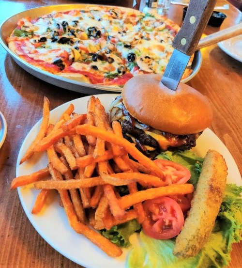 A plate with a burger topped with cheese, sweet potato fries, lettuce, and tomato, with a pizza in the background.