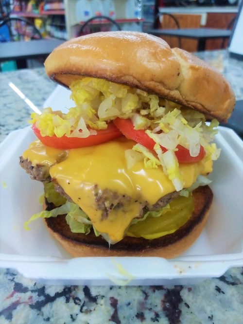 A cheeseburger with lettuce, tomato, pickles, and a toasted bun, served on a white takeout container.