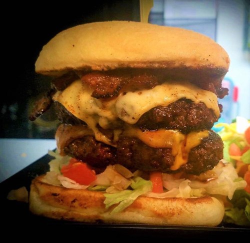A towering burger with two beef patties, melted cheese, and toppings, served on a toasted bun with lettuce and tomato.