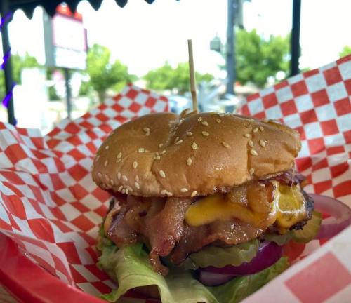 A sesame seed bun burger with bacon, cheese, pickles, and lettuce on a red and white checkered plate.