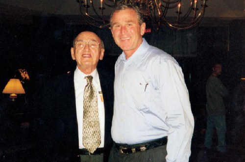 Two men smiling together in a well-lit room, with a chandelier and soft lighting in the background.