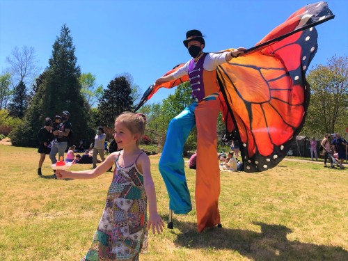 A child in a colorful dress holds ice cream while a stilt walker in a butterfly costume poses nearby in a sunny park.