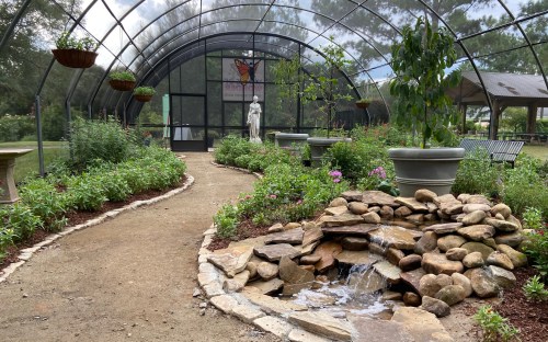 A serene garden path leads to a greenhouse, featuring a small stone waterfall and lush greenery.