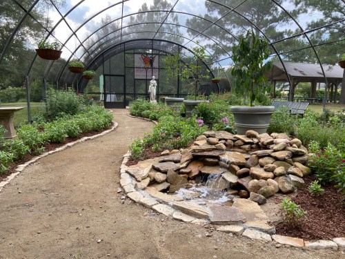 A serene garden path with a stone waterfall, surrounded by lush greenery and a greenhouse structure in the background.