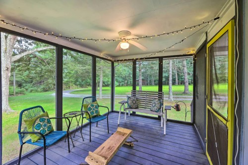 A screened porch with two chairs, a bench, and string lights, surrounded by greenery and trees.