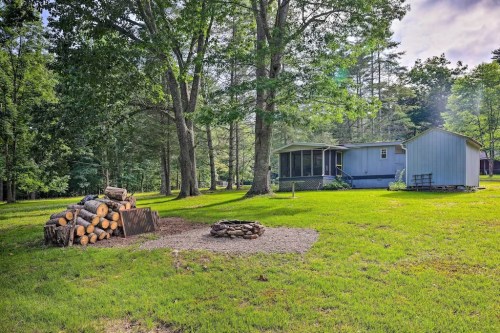 A grassy yard with a fire pit, stacked firewood, and a blue shed surrounded by trees and a house in the background.