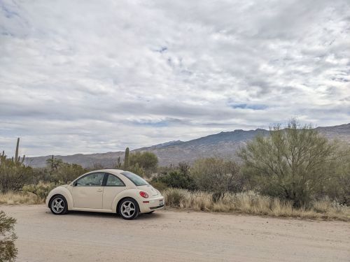 A beige Volkswagen Beetle parked on a dirt road with mountains and desert vegetation in the background under a cloudy sky.