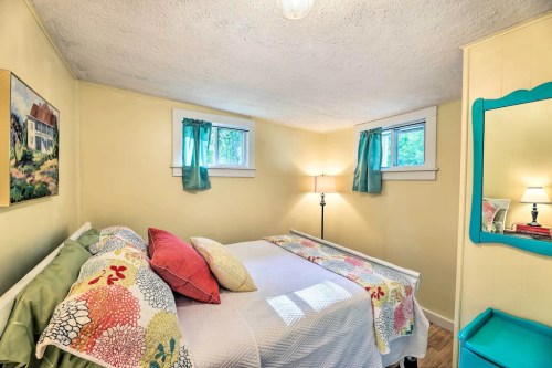 Cozy bedroom with a double bed, colorful bedding, two windows, and a lamp, featuring light yellow walls and a painting.