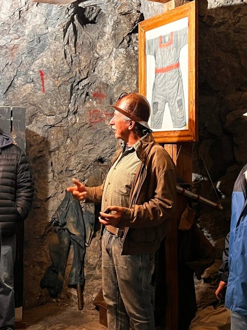 A man in a hard hat speaks in a rocky underground setting, with a framed illustration of work gear on the wall behind him.