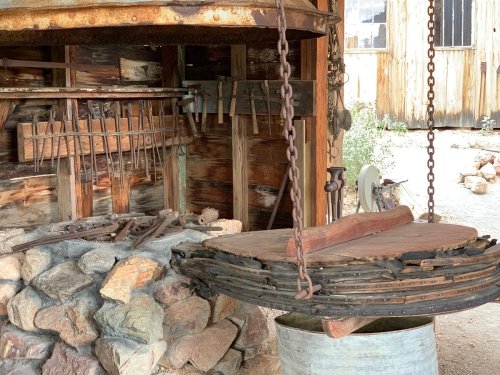 An old wooden scale hangs over a stone base, surrounded by rustic tools and weathered wooden structures.