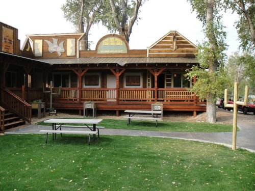 A rustic wooden building with a porch, surrounded by trees and grassy areas, featuring picnic tables in front.