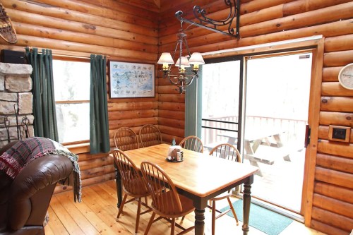 Cozy log cabin dining area with a wooden table, chairs, and a chandelier, featuring large windows and a sliding door.