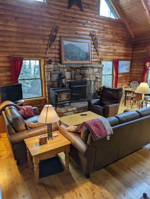 Cozy log cabin interior with a stone fireplace, wooden furniture, and large windows letting in natural light.