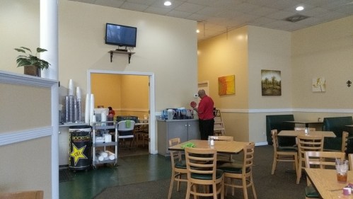 A spacious restaurant interior with tables, a TV on the wall, and a staff member preparing drinks at a counter.