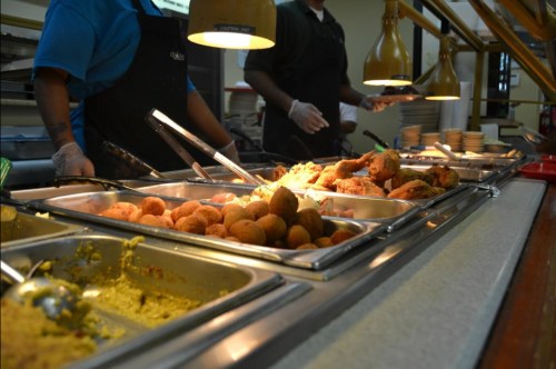 A buffet line with various dishes, including fried food and sides, being served by staff in a cafeteria setting.