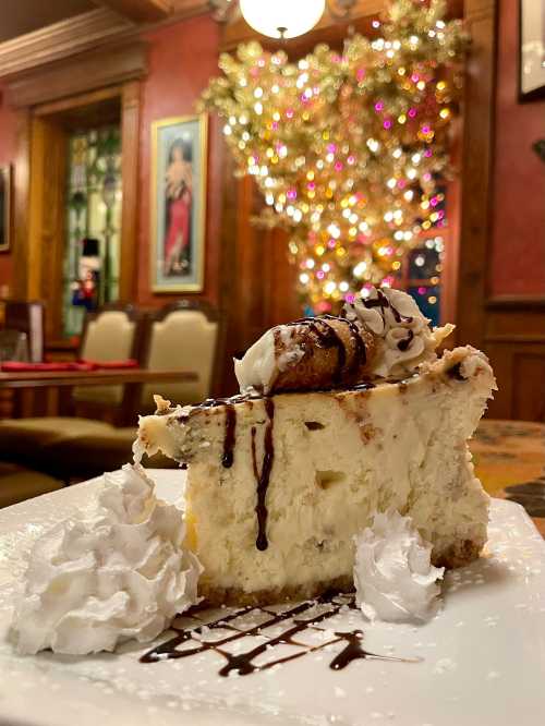 A slice of cheesecake topped with whipped cream and chocolate drizzle, served on a plate with festive decor in the background.