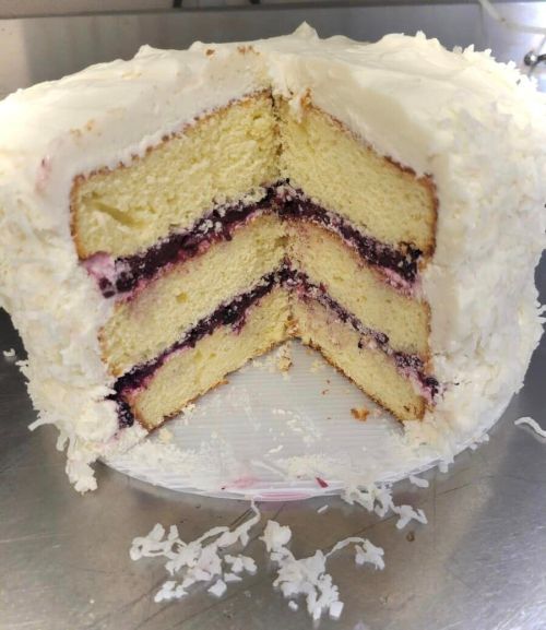 A sliced cake with layers of yellow sponge and purple filling, topped with white frosting and coconut flakes.