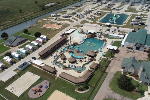 Aerial view of a resort with a large pool, cabanas, and RV parking area, surrounded by green fields and water.