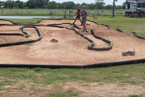 Two people stand by a dirt track with remote-controlled cars racing on it, surrounded by greenery and a pond.
