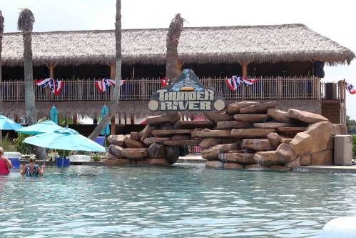 A tropical pool area featuring a rock formation and a sign for "Thunder River," with a thatched-roof building in the background.