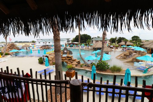 A tropical resort pool area with palm trees, cabanas, and turquoise umbrellas, surrounded by lush landscaping.