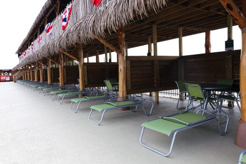 A row of empty lounge chairs and tables under a thatched roof, decorated with red, white, and blue banners.