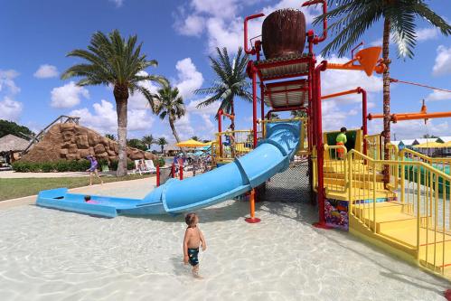 A child stands in shallow water near a colorful water slide and play structure, surrounded by palm trees and sunny skies.