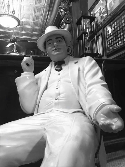 A life-sized statue of a man in a white suit and hat, seated with a relaxed pose in a vintage setting.