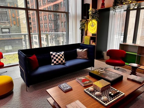 A modern living room with a navy sofa, colorful chairs, a coffee table, and large windows overlooking a cityscape.