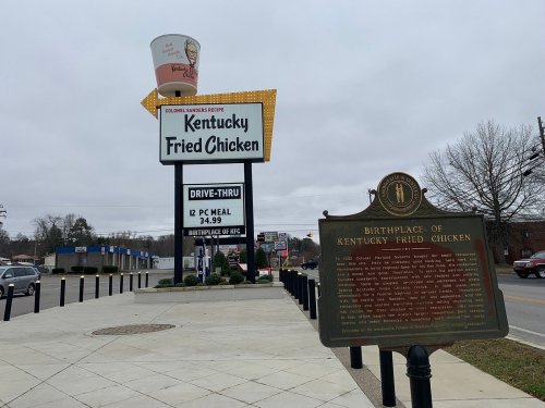Sign for Kentucky Fried Chicken with a historical marker about its birthplace, set against a cloudy sky.