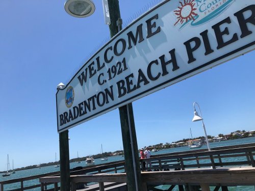 Sign welcoming visitors to Bradenton Beach Pier, established in 1921, with boats visible in the background.