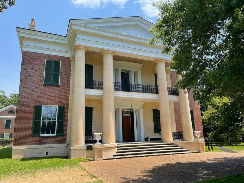 A grand two-story brick mansion with large columns, green shutters, and a wide staircase, surrounded by trees.