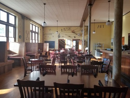 A spacious restaurant with wooden tables and chairs, featuring artistic wall decor and large windows.