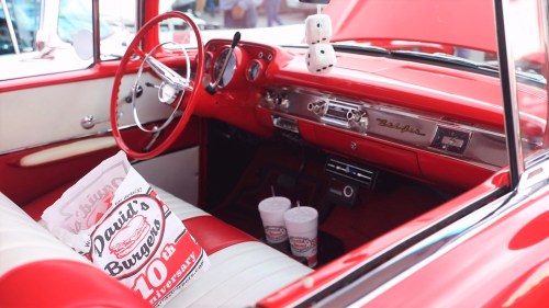 Interior of a vintage red car featuring white leather seats, a classic dashboard, and takeout bags from David's Burgers.