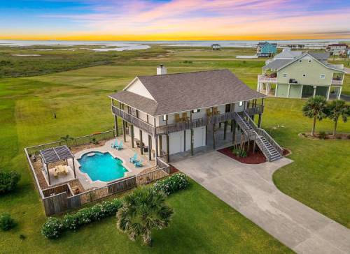 Aerial view of a beach house with a pool, surrounded by green grass and a colorful sunset in the background.