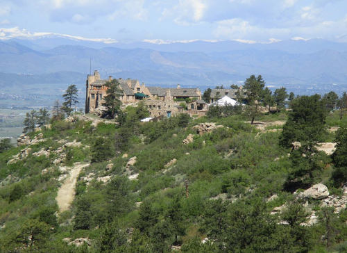 A stone building on a hillside surrounded by trees, with mountains and a blue sky in the background.