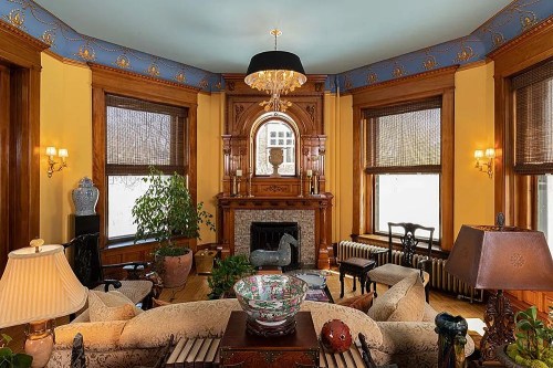 Cozy living room with ornate woodwork, a chandelier, and large windows, featuring plants and decorative furnishings.