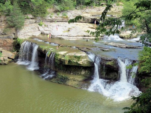 A scenic view of waterfalls cascading over rocks into a calm river, surrounded by lush greenery and people enjoying the area.