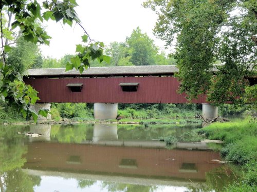 A red covered bridge spans a calm river, surrounded by lush green trees and reflecting in the water below.