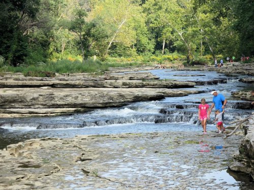 A family walks along a rocky riverbank, enjoying a sunny day by the water with lush greenery in the background.