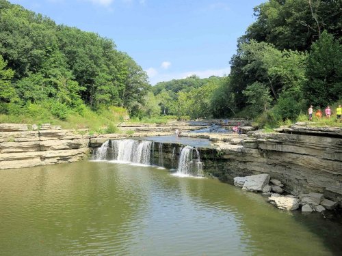 A serene landscape featuring a waterfall cascading into a calm pool, surrounded by lush greenery and rocky cliffs.