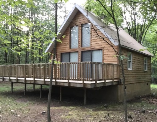 A wooden cabin with large windows and a deck, surrounded by trees in a forested area.