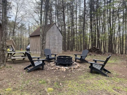 A cozy outdoor fire pit area with black Adirondack chairs surrounded by trees and a wooden shed in the background.