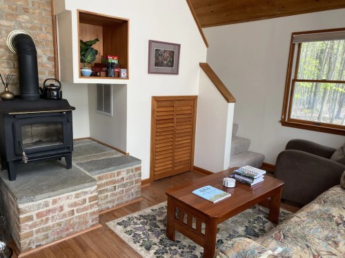 Cozy living room with a wood stove, wooden coffee table, and a staircase leading to an upper level. Natural light from a window.