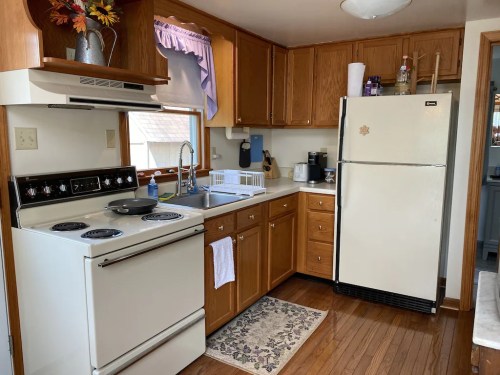 A cozy kitchen with wooden cabinets, a white stove, sink, and refrigerator, featuring a rug and floral decor.