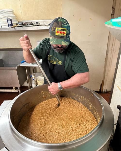 A person in a camo hat stirs a large pot of food in a kitchen setting.